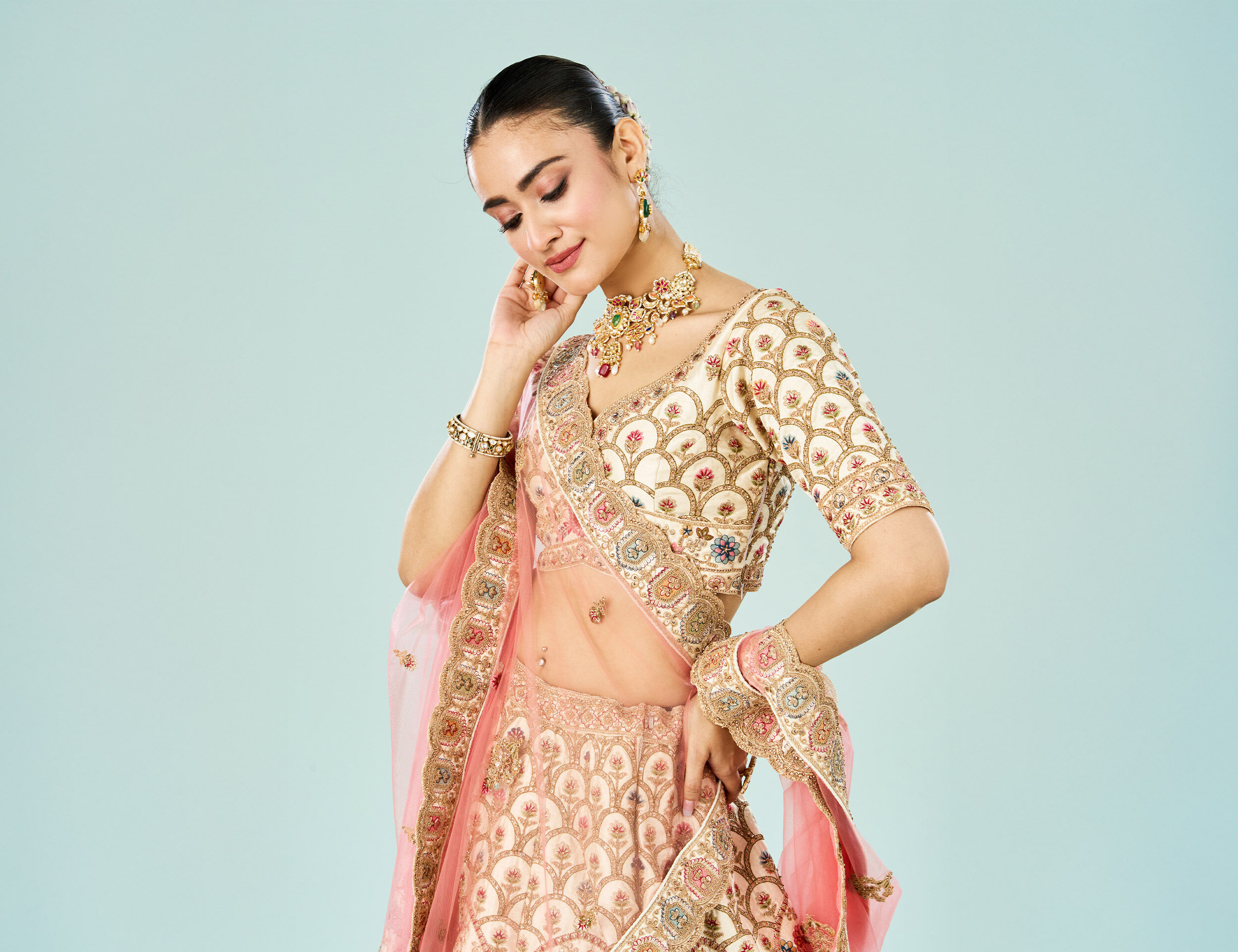 Bride in a pastel peach lehenga with intricate gold and maroon embroidery, matching blouse and dupatta, styled with floral jewelry against a light blue background